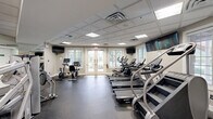 Located at 1 Applewood Drive in Perkasie, Pennsylvania, this fitness center offers a generously sized, single-floor exercise space designed to support a comprehensive workout experience. The room features a drop tile ceiling with recessed and flush-panel lighting that ensures even, bright illumination throughout the space.

The facility is well-appointed with a diverse array of equipment. The cardio zone includes multiple treadmills by Spirit, a stair climber, elliptical trainers, and stationary bikes, providing ample options for aerobic exercise. The strength training area is equipped with a Hoist multi-functional cable machine, a leg press machine, a weight bench, and a cable cross machine, along with a medicine ball rack for functional fitness training.

Durable rubber flooring covers the main exercise areas, transitioning to tile near the entryway. Large windows with blinds along one wall, flanked by French doors, allow natural light to fill the space while offering views of the surrounding greenery. Wall-mounted flat-screen TVs are positioned throughout for entertainment during workouts. Full-length mirrors on one wall enhance the sense of openness and allow for form monitoring. The neutral gray and beige color palette, combined with a pass-through window to an adjacent room, gives the facility a clean, professional, and welcoming atmosphere.