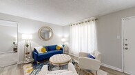 This single-floor residence offers a thoughtfully designed layout that balances comfort and functionality across its key living areas. The living room serves as the heart of the home, featuring wood-look flooring, a textured ceiling, and a large window dressed with sheer grommet-panel curtains that fill the space with natural light. A front entry door opens directly into this area, which flows toward a dedicated dining space separated by a stepped ceiling detail and connected to the kitchen via a pass-through counter.

The galley-style kitchen is equipped with gray cabinetry, light stone-look countertops, a stainless steel sink, and a full suite of black and stainless steel appliances, including a range, microwave, refrigerator, and dishwasher. The adjacent dining area comfortably accommodates a round table with multiple seats and features wood-look flooring consistent with the main living areas.

Both bedrooms are carpeted and include ceiling fans with light fixtures and windows with curtain panels. Each room offers generous space for a full bedroom suite. The two bathrooms mirror each other in design, each featuring gray vanity cabinetry, stone-look countertops, a single sink, and a tub-shower combination with tile surround.