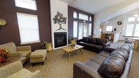 This single-floor clubhouse offers a warm and inviting gathering space designed for comfort and community. The main lounge area immediately draws attention with its soaring vaulted ceilings and dramatic two-tone walls, featuring rich deep plum accent walls contrasted by crisp white surfaces that enhance the sense of height and openness. A central fireplace with a sleek black surround serves as the focal point of the room, framed by a striking metal tree wall sculpture that adds an artistic touch to the space. Natural light pours in through multiple windows fitted with white blinds, creating a bright yet cozy atmosphere throughout the day. The lounge is furnished with dark leather sofas and upholstered accent chairs paired with an ottoman, all arranged around a mid-century modern wooden coffee table atop a patterned area rug. Lush potted plants add a refreshing natural element to the décor. A glimpse into an adjacent area reveals a kitchenette, suggesting additional amenities available for gatherings and events. The overall ambiance of the clubhouse is both stylish and functional, making it a versatile space well-suited for casual lounging, social events, and community activities.