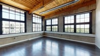 This single-floor loft space masterfully blends industrial character with modern finishes, creating a striking and versatile interior. The open main living area is defined by soaring wood-plank ceilings supported by heavy timber beams, exposed metal ductwork, and original concrete walls that speak to the building's heritage. Dark-stained hardwood floors run throughout, reflecting the abundant natural light that pours in through an impressive array of large, black steel-framed multi-pane windows. These oversized windows offer expansive views of the surrounding cityscape and greenery, lending the space an airy, gallery-like quality.

The kitchen is outfitted with contemporary two-tone cabinetry, white quartz countertops, a white tile backsplash, and stainless steel appliances, offering a clean, modern contrast to the raw industrial backdrop. A private room features the same warm hardwood flooring and carries the timber beam ceiling detail, maintaining design continuity throughout. The bathroom is finished with a glass-enclosed walk-in shower, modern vanity with white countertop, and wood-look tile flooring, providing a bright and functional space. Together, these elements create a cohesive loft environment that balances raw architectural character with refined, contemporary convenience.
