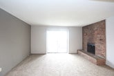 This single-floor residence offers a practical and straightforward layout designed for comfortable everyday living. The living area is finished with wall-to-wall beige carpeting and features a textured ceiling, a wall-mounted light fixture, and access to adjacent rooms through dark-stained wood doors. Natural light enters through a window, lending the space a bright, open feel.

The kitchen is positioned at the heart of the unit and features a peninsula-style countertop with a built-in stainless steel sink, creating a pass-through connection to the living area. Dark wood upper cabinetry provides ample storage, while white appliances — including a refrigerator and an electric range — complete the functional cooking space.

The bedroom continues the carpeted flooring found throughout, offering a clean, neutral environment. A window with horizontal blinds allows natural light in while maintaining privacy. The textured ceiling and flush-mount light fixture provide consistent finishing details across rooms.

The bathroom includes warm-toned hardwood flooring, a dark wood vanity with a light-colored countertop and integrated sink, and a wall-mounted towel bar. The space is efficiently organized, reflecting a no-frills, utilitarian design. Overall, the unit presents a cohesive, understated aesthetic suited for those seeking a functional and low-maintenance living space.