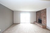 This single-floor residence offers a practical and straightforward layout designed for comfortable everyday living. The living area is finished with wall-to-wall beige carpeting and features a textured ceiling, a wall-mounted light fixture, and access to adjacent rooms through dark-stained wood doors. Natural light enters through a window, lending the space a bright, open feel.

The kitchen is positioned at the heart of the unit and features a peninsula-style countertop with a built-in stainless steel sink, creating a pass-through connection to the living area. Dark wood upper cabinetry provides ample storage, while white appliances — including a refrigerator and an electric range — complete the functional cooking space.

The bedroom continues the carpeted flooring found throughout, offering a clean, neutral environment. A window with horizontal blinds allows natural light in while maintaining privacy. The textured ceiling and flush-mount light fixture provide consistent finishing details across rooms.

The bathroom includes warm-toned hardwood flooring, a dark wood vanity with a light-colored countertop and integrated sink, and a wall-mounted towel bar. The space is efficiently organized, reflecting a no-frills, utilitarian design. Overall, the unit presents a cohesive, understated aesthetic suited for those seeking a functional and low-maintenance living space.