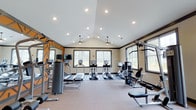 Located at 45 Lullwater Dr in Richmond Hill, Georgia, this single-floor fitness center is designed to support a well-rounded workout experience. The space features vaulted ceilings that lend a sense of openness and volume, complemented by recessed lighting and ceiling fans that ensure a comfortable environment during exercise. Multiple large windows line the walls, flooding the interior with natural light and offering views of the surrounding wooded landscape. The equipment layout is thoughtfully organized, with a dedicated cardio zone housing treadmills and elliptical trainers positioned near the windows, alongside a multi-station strength training machine and additional resistance equipment. A wall-mounted flat-screen television adds convenience for entertainment during workouts. The accent wall features a bold graphic detail, adding an energetic visual element to the space. The neutral color palette, clean flooring, and dark trim details give the facility a polished, modern aesthetic. This fitness center provides residents with a well-equipped, inviting space for their daily exercise routines, all set within a bright, airy environment that makes working out an enjoyable experience.