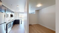 This single-floor unit offers a clean, modern design highlighted by light hardwood-style flooring that runs throughout the main living areas. The open-concept kitchen seamlessly connects to the living space, creating a bright and functional layout. The kitchen is well-appointed with rich dark cabinetry, granite countertops, stainless steel appliances including a refrigerator, range, microwave, and dishwasher, and a double-basin sink. Stylish pendant lighting hangs above the kitchen peninsula, which provides additional counter space and serves as a casual dining area. Large windows and a sliding glass door open to a private balcony, flooding the living area with natural light and offering pleasant outdoor views. A dedicated bedroom area features neutral-toned walls and ample space, with access to a closet equipped with a hanging rod. The bathroom is finished with earth-toned tile flooring, a dark wood vanity with a white countertop, a wall-mounted mirror, and a shower-tub combination. Clean white walls, flush-mount ceiling fixtures, and baseboard heating throughout contribute to the unit's consistent, understated aesthetic.