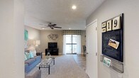 This single-floor residence offers a welcoming and functional layout centered around a spacious living area. The living room is finished with plush wall-to-wall carpeting that transitions to smooth wood-look hard flooring near the entryway, creating a natural distinction between zones. Neutral taupe walls provide a warm, versatile backdrop throughout the space, complemented by a textured popcorn ceiling with recessed lighting and a ceiling fan that enhances both comfort and ambiance.

Natural light flows generously into the room through a large sliding glass door framed by sheer white curtains and a decorative teal-patterned valance, offering a view of and direct access to an outdoor patio area. The living room comfortably accommodates a seating arrangement anchored by a glass-top coffee table, with ample space for media furniture. Near the entryway, a chalkboard wall organizer adds a practical, personalized touch. The overall atmosphere is casual yet polished, with thoughtful design details that balance everyday livability with a tidy, modern aesthetic.