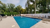 Located on Lighthouse Lane in East Lyme, Connecticut, this outdoor pool facility offers a well-appointed recreational retreat designed for both relaxation and leisure. The centerpiece of the space is a large, rectangular swimming pool with crystal-clear blue water, accessible via built-in entry steps and handrails. The pool is surrounded by an expansive concrete sundeck that provides ample space for lounging, lined on all sides with rows of sling-style chaise lounges positioned to take advantage of the open sky and natural light.

The perimeter of the facility is enclosed by a combination of black aluminum fencing and white vinyl fencing, offering both security and a clean, polished aesthetic. Neatly trimmed arborvitae hedges and mature deciduous trees frame the space, creating a natural, private backdrop that enhances the serene atmosphere.

A charming single-story poolhouse anchors one end of the facility, featuring a classic New England-style pitched roof with a cupola, wide windows, and a covered porch area outfitted with outdoor dining tables and chairs shaded by blue market umbrellas — ideal for casual gatherings. Residential buildings are visible beyond the fence line, situating this facility as a shared amenity within a broader community. The overall design balances functionality with comfort, making it an inviting outdoor destination throughout the warmer months.