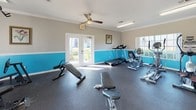 This single-floor fitness center offers a comprehensive workout environment designed to accommodate a variety of exercise routines. The space is immediately distinguished by its two-tone wall treatment, featuring a bold turquoise lower half paired with a neutral taupe upper half, accented by white crown molding that adds a polished finishing touch. Durable dark rubber flooring covers the entire area, providing a safe and practical surface for all types of physical activity.

The cardio section is well-appointed with a treadmill, recumbent bike, upright stationary bike, and an elliptical trainer, all positioned near windows that allow natural light to fill the space and offer views of the surrounding greenery. A wall-mounted flat-screen television is conveniently positioned for entertainment during workouts. The strength and free weight area features a multi-tiered dumbbell rack stocked with a full range of weights, accompanied by adjustable workout benches and additional strength training machines.

Ceiling fans help maintain a comfortable temperature throughout the facility. Multiple sets of French doors on opposing walls promote excellent ventilation and enhance the open, airy feel of the room. A hand sanitizer dispenser and small storage unit are also present, adding to the practicality of the space. Ample natural light from multiple windows completes this inviting and functional fitness environment.