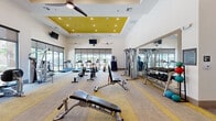 This modern fitness center offers a generously sized, single-floor layout designed to accommodate a comprehensive range of workout routines. The space is defined by soaring ceilings with a distinctive coffered section finished in bold yellow, adding a vibrant, energetic accent to the otherwise clean white interior. Multiple ceiling fans and recessed lighting keep the environment comfortable and well-lit throughout.

Large floor-to-ceiling windows line the perimeter walls, flooding the main workout area with natural light and providing views of the surrounding outdoor greenery. The primary cardio zone features a row of treadmills, stationary bikes, rowing machines, ellipticals, and a stair climber, all positioned to take advantage of the natural light and outdoor views.

The strength training section is equipped with a multi-functional cable machine, a full dumbbell rack, and multiple adjustable benches, complemented by wall-to-wall mirrors that enhance the sense of space. A dedicated functional training and stretching area features hardwood-style flooring, large mirrors, stability balls, and medicine balls, providing a versatile space for yoga, stretching, or group exercise.

The flooring throughout transitions thoughtfully between patterned carpet in the main cardio and strength areas and smooth hardwood in the functional zone. A bulletin board, entry doors with glass panels, and a storage area round out the practical amenities of this well-appointed fitness facility.