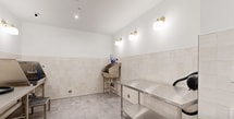 Located at 6 Becker Farm Rd in Roseland, NJ, this 180-square-foot dedicated pet grooming room is a well-appointed, single-floor facility designed for functionality and ease of use. The room features neutral white upper walls with cream-toned square tile wainscoting along the lower half, creating a clean and hygienic aesthetic throughout. Large-format stone-look porcelain floor tiles add durability and a polished appearance underfoot, with centrally placed floor drains for efficient water management. The space is equipped with multiple stainless steel grooming tables positioned along the perimeter walls, a deep stainless steel wash tub with a coiled blue hose for bathing, and professional-grade pet dryers mounted at each grooming station. Brass-finish wall sconces provide warm, even lighting throughout the room, complemented by recessed ceiling fixtures. A sturdy wooden bench near the entry door offers a convenient seating area, accompanied by posted WAG facility rules for user reference. The heavy-duty steel entry door with a window panel ensures secure access. Smoke and fire detection equipment is also present, reflecting a commitment to safety. This thoughtfully designed grooming space delivers a complete, professional-grade environment for pet care.