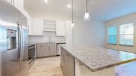 This single-floor residence showcases a thoughtfully designed open-concept layout that seamlessly connects the kitchen and living areas. The kitchen serves as the focal point, featuring a large granite-topped island with built-in storage and pendant lighting overhead. White upper cabinetry contrasts with warm wood-toned lower cabinets, while a white subway tile backsplash adds a clean, contemporary detail. A full suite of stainless steel appliances includes a side-by-side refrigerator with ice and water dispenser, electric range, built-in microwave, and dishwasher. Light hardwood-style flooring extends throughout the main living areas, contributing to a bright and cohesive aesthetic. Large windows fitted with horizontal blinds allow natural light to filter through the living space, which also features access to an outdoor area via a glass door. The bedroom areas offer plush carpeting in at least one room and hardwood-style flooring in another, each with windows providing natural light and outdoor views. The overall design palette is neutral and contemporary, emphasizing clean lines, modern finishes, and a bright, airy atmosphere throughout.