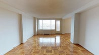 This single-floor space is characterized by its open layout and an abundance of natural light streaming through a large picture window at the far end of the main living area. The window is flanked by a glass-paneled door that provides direct access to a balcony, offering views of the surrounding neighborhood. The spacious main living area is finished with glossy parquet hardwood flooring in a warm honey tone, arranged in a classic block pattern that adds visual texture and a timeless aesthetic to the space. The walls are painted a clean, neutral white, complementing the light-colored flooring and enhancing the bright, airy atmosphere throughout. A textured popcorn ceiling adds a subtle architectural detail overhead. A baseboard heating unit is positioned beneath the picture window, ensuring consistent warmth throughout the colder months. The open floor plan allows for flexible furniture arrangements and seamless flow between living zones. The overall ambiance is clean, uncluttered, and well-lit, making it a versatile space suitable for a variety of uses and interior design styles.