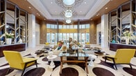 Located at 1517 Cameron Samuel Ln in West Palm Beach, Florida, this grand lobby space delivers a striking first impression through its contemporary design and carefully curated interiors. The entry corridor features bold striped marble-and-tile flooring in cream and dark brown tones, flanked by a sculptural interlocking-circle ceiling detail that adds architectural depth and visual interest. A sleek marble reception desk with warm wood accents anchors the entry area, offering a polished and professional welcome.

The space opens into a dramatically scaled lounge area crowned by soaring coffered ceilings with recessed lighting and a layered, ornate chandelier that serves as a stunning focal point. Floor-to-ceiling windows and glass doors flood the interior with abundant natural light while framing views of the lush Florida landscape beyond. The lounge is furnished with crisp white sofas accented by vibrant teal and yellow throw pillows, arranged atop a bold circular-patterned area rug in cream and espresso brown.

Decorative room dividers with geometric lattice-cut panels define zones within the open plan, while large-scale abstract artwork and curated sculptural accents add cultural richness. Warm wood wall paneling contrasts beautifully with the bright white palette, lending a refined, resort-like ambiance throughout this thoughtfully designed West Palm Beach property.