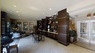 This single-floor clubroom offers a thoughtfully designed communal space that balances style and functionality. The room is anchored by a prominent dark walnut-toned built-in shelving unit that spans a significant portion of the wall, featuring open display shelves adorned with decorative plants, books, and curated accessories, alongside lower cabinet storage. Recessed lighting throughout the ceiling ensures the space is bright and welcoming, complementing the light-toned tile flooring that runs throughout. A casual dining area with bistro-style metal chairs and small tables is positioned near the shelving wall, providing a relaxed spot for informal gatherings or individual use. Adjacent to this, a lounge area is furnished with a navy sofa, woven accent chairs, and a low coffee table, creating an inviting setting for socializing or unwinding. Tall potted plants and a sculptural wall accent add natural and artistic touches, enhancing the room's contemporary ambiance. Large framed mirrors along one wall reflect light and contribute to a sense of openness. A wall-mounted television integrated into the shelving unit adds a media-friendly element to the space. Overall, the clubroom presents a polished, modern environment well-suited for both casual relaxation and community interaction.