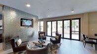 This well-appointed indoor space combines functionality and contemporary design across a single floor. The lounge area serves as a central highlight, featuring a sophisticated seating arrangement with dark leather armchairs, a patterned area rug, and a wooden coffee table accented with decorative accessories. A striking textured accent wall in a metallic, geometric finish anchors the room and provides a stylish backdrop for the wall-mounted flat-screen television. Recessed lighting in the white ceiling illuminates the space with a clean, modern feel. One of the most notable features is the expansive floor-to-ceiling glass sliding doors that line the exterior wall, flooding the interior with abundant natural light and offering open views of the surrounding outdoor greenery and adjacent buildings. The seamless connection between the interior and exterior creates an airy, open ambiance. The overall design aesthetic is polished and contemporary, with a neutral color palette of whites, grays, and warm browns that lends a welcoming yet refined atmosphere to the space — ideal for gathering, relaxing, or entertaining.