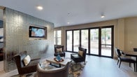 This well-appointed indoor space combines functionality and contemporary design across a single floor. The lounge area serves as a central highlight, featuring a sophisticated seating arrangement with dark leather armchairs, a patterned area rug, and a wooden coffee table accented with decorative accessories. A striking textured accent wall in a metallic, geometric finish anchors the room and provides a stylish backdrop for the wall-mounted flat-screen television. Recessed lighting in the white ceiling illuminates the space with a clean, modern feel. One of the most notable features is the expansive floor-to-ceiling glass sliding doors that line the exterior wall, flooding the interior with abundant natural light and offering open views of the surrounding outdoor greenery and adjacent buildings. The seamless connection between the interior and exterior creates an airy, open ambiance. The overall design aesthetic is polished and contemporary, with a neutral color palette of whites, grays, and warm browns that lends a welcoming yet refined atmosphere to the space — ideal for gathering, relaxing, or entertaining.