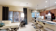 This single-floor residence in Dallas offers a thoughtfully designed open-concept layout that seamlessly connects the living and kitchen areas, creating a bright and inviting atmosphere. The living space is anchored by large windows dressed with wood-toned blinds and elegant floor-to-ceiling drapes in a bold charcoal hue, allowing natural light to fill the room while providing privacy. A chic accent floor lamp and patterned area rug add warmth and character to the living area, which features clean white tile flooring throughout.

The kitchen is a standout feature, showcasing rich warm-toned cabinetry, a curved breakfast bar with modern metallic counter stools, and a pendant light that lends a contemporary touch. A second dining nook is visible near the window, complete with additional seating and decorative valances in a teal and neutral pattern that ties the color palette together. The kitchen also offers generous counter space and a functional layout ideal for both casual dining and entertaining.

The overall aesthetic blends modern and transitional design elements, with a neutral base accented by teal and dark tones throughout. The space feels open and airy, with natural light playing a key role in highlighting the thoughtful interior details.