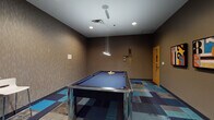 This single-level game room is thoughtfully designed as a dedicated entertainment space with a contemporary aesthetic. The room is anchored by two key gaming features: a full-size billiards table with a blue felt surface and a metallic base, positioned beneath a sleek multi-pendant light fixture, and a large oval poker table with a blue felt top, black leather padded rail, and built-in cup holders surrounded by cushioned rolling chairs. A wall-mounted cue rack keeps billiards accessories organized and accessible. The poker area is equipped with a wall-mounted flat-screen television and adjacent cabinetry, adding functional convenience to the space. A compact bistro table with two white chairs occupies a corner, offering a casual seating option. The flooring features a patchwork carpet design in shades of blue, teal, gray, and navy, contributing a bold visual element throughout. Warm taupe textured wall coverings line the room, complemented by recessed lighting and pendant fixtures that provide focused illumination. A vibrant abstract wall painting adds a striking artistic focal point. The amber-toned entry door and framed graphic artwork on the walls complete the modern, curated atmosphere of this well-appointed game room.