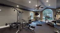 This fitness center at 1000 Glen Forest Dr in Greenville, SC is a well-appointed, single-story facility designed to support a comprehensive workout experience. The space features vaulted ceilings that lend an open, airy feel, complemented by recessed lighting and a ceiling fan that enhance both visibility and comfort during exercise.

Natural light flows generously through multiple windows, including a prominent arched window with a decorative grid pattern that serves as a striking architectural focal point. The view of mature surrounding greenery adds a pleasant outdoor connection to the interior environment.

The flooring throughout is durable rubber gym matting, practical for both cardio and strength training use. Full-length mirrored walls on one side visually expand the space while providing functional feedback for users during workouts.

Equipment is well-distributed across the floor and includes multiple treadmills, an elliptical trainer, a stationary bike, a multi-station cable machine, weight benches, and a dumbbell rack. A wall-mounted television is positioned for easy viewing from cardio equipment.

A glass-paneled door provides direct exterior access to the surrounding landscaped grounds. The neutral gray wall tones and thoughtful layout create a clean, professional atmosphere suited for focused fitness activity.