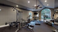 This fitness center at 1000 Glen Forest Dr in Greenville, SC is a well-appointed, single-story facility designed to support a comprehensive workout experience. The space features vaulted ceilings that lend an open, airy feel, complemented by recessed lighting and a ceiling fan that enhance both visibility and comfort during exercise.

Natural light flows generously through multiple windows, including a prominent arched window with a decorative grid pattern that serves as a striking architectural focal point. The view of mature surrounding greenery adds a pleasant outdoor connection to the interior environment.

The flooring throughout is durable rubber gym matting, practical for both cardio and strength training use. Full-length mirrored walls on one side visually expand the space while providing functional feedback for users during workouts.

Equipment is well-distributed across the floor and includes multiple treadmills, an elliptical trainer, a stationary bike, a multi-station cable machine, weight benches, and a dumbbell rack. A wall-mounted television is positioned for easy viewing from cardio equipment.

A glass-paneled door provides direct exterior access to the surrounding landscaped grounds. The neutral gray wall tones and thoughtful layout create a clean, professional atmosphere suited for focused fitness activity.
