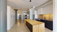 This single-floor loft in Memphis, TN offers a thoughtfully designed open-concept layout that blends industrial character with modern finishes. Polished concrete floors run throughout the space, complementing the exposed ductwork and structural columns that lend an authentic loft aesthetic. The kitchen is a standout feature, showcasing dark lower cabinetry, white upper cabinets, a warm wood-toned backsplash, stainless steel appliances including a refrigerator and range, and a generous butcher block island that doubles as a prep and dining surface. A sleek linear pendant light anchors the kitchen area. Adjacent to the kitchen, a glass-panel partition with black metal framing encloses a stacked in-unit washer and dryer — a practical and visually striking design element. The open living area benefits from natural light provided by a large window, keeping the white walls bright and airy. The full bathroom features polished concrete flooring, a frameless sliding glass walk-in shower with white tile surround, a vanity with storage, and clean, minimalist finishes throughout. The entry door is a bold slate gray with matte black hardware, setting the tone for the space's contemporary industrial design.