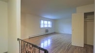 This single-floor space offers a straightforward and functional layout with a number of noteworthy details. The main living area is anchored by polished hardwood floors that reflect natural light beautifully, giving the space a warm and inviting feel. A multi-pane window along the exterior wall allows natural light to filter in, brightening the room and creating an airy atmosphere. Baseboard heating runs along the perimeter of the living area, ensuring efficient and even warmth throughout. A wall-mounted air conditioning unit beneath the window provides climate control year-round. The neutral, light-toned walls offer a clean, versatile backdrop suitable for a variety of interior styles. A decorative wrought iron staircase railing with ornate scroll detailing adds a classic architectural touch and serves as a visual focal point within the space. The overall layout is open and unobstructed, maximizing usable floor space. A doorway visible along the side wall suggests additional rooms or access points beyond the main area. The combination of classic hardwood flooring, practical heating and cooling solutions, and thoughtful architectural accents makes this a well-rounded and comfortable space.
