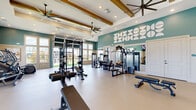 This fitness center offers a comprehensive workout environment across a generously sized single-floor layout, seamlessly combining indoor and outdoor training spaces. The interior is defined by elevated ceilings with exposed wood beam accents, recessed lighting, and large ceiling fans that contribute to an open, airy atmosphere. Teal-painted upper walls paired with crisp white wainscoting create a vibrant yet clean aesthetic, reinforced by geometric wall art and motivational signage.

The indoor area is divided into functional training zones. A strength section houses cable machines, multi-functional racks, weight benches, and a full dumbbell rack. A dedicated cardio zone features treadmills, spin bikes, and an air bike, positioned near floor-to-ceiling windows that flood the space with natural light and offer views of the surrounding palm-lined landscape. Full-length mirrors and a wall-mounted display screen further enhance usability.

A roll-up garage door opens directly to the outdoor turf training area, which includes a pull-up rig, barbell stations, plyo boxes, kettlebells, and a storage cabinet for additional equipment. The outdoor space is enclosed by iron fencing, providing a secure and functional open-air training environment.