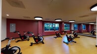 This indoor cycling studio is a purpose-built fitness space designed to support focused, high-energy workouts. The room is outfitted with a fleet of stationary cycling bikes arranged in rows across a smooth, light-toned hardwood-style floor that provides both durability and a clean aesthetic. Bold deep-red accent walls contrast with neutral white surfaces, creating an energizing atmosphere suited to intense exercise sessions.

Overhead, large circular flush-mount light fixtures provide bright, even illumination throughout the space. A full-width wall mirror at one end of the room enhances the sense of depth while allowing participants to monitor their form during workouts. A wall-mounted flat-screen television is positioned for visibility across the room, ideal for instructor-led classes or on-demand cycling programs.

Natural light filters in through a row of windows along one wall, offering views of outdoor greenery and adding a refreshing visual element to the environment. A dedicated storage rack holds a variety of exercise balls, including medicine balls and stability balls, providing supplemental training options. The studio is accessed through a set of glass-paneled double doors, which connect the space to adjacent areas of the facility. The overall layout is open and well-organized, supporting both group classes and individual training sessions.