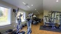 This fitness center is a dedicated, single-floor workout facility designed to support a comprehensive exercise routine. The space is outfitted with a variety of cardio equipment, including multiple elliptical trainers and stationary bikes, arranged to maximize usability and flow throughout the room. Strength and resistance training equipment lines the walls, offering a well-rounded selection for users of all fitness levels. The flooring consists of warm hardwood, complemented by rubber exercise mats in designated workout zones, providing both durability and comfort underfoot. Large windows along one wall invite generous natural light, creating a bright and energizing atmosphere that enhances the workout experience. Recessed lighting in the ceiling ensures consistent, even illumination throughout the space. The walls are finished in a neutral, light tone that contributes to the clean and open feel of the room. Ventilation is supported by ceiling vents, keeping the environment comfortable during intense workouts. Overall, the fitness center offers a practical, well-maintained environment suited for daily exercise, combining functional equipment layout with an inviting, light-filled ambiance.