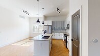 This well-appointed single-floor residence offers a thoughtfully designed layout that balances modern aesthetics with everyday functionality. The open-concept kitchen and living area serves as the heart of the home, showcasing striking herringbone-patterned hardwood flooring that flows seamlessly throughout the main living spaces. The kitchen is outfitted with a mix of crisp white and soft gray shaker-style cabinetry, complemented by matte black hardware, a marble-inspired subway tile backsplash, and light quartz countertops. A central island with a dark countertop and an undermount sink anchors the space, while pendant lighting overhead adds a contemporary touch. Stainless steel appliances complete the culinary setup. The bedrooms feature plush wall-to-wall carpeting and ceiling fans, with several rooms benefiting from multiple windows that invite generous natural light. The bathrooms are finished to a high standard, featuring white shaker vanities with dark countertops, backlit LED mirrors, matte black fixtures, large-format tile flooring, and a glass-enclosed walk-in shower in the primary bath. The overall ambiance of the space is clean, bright, and modern, with a neutral color palette that creates a welcoming and versatile living environment.