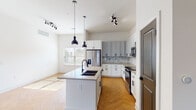 This well-appointed single-floor residence offers a thoughtfully designed layout that balances modern aesthetics with everyday functionality. The open-concept kitchen and living area serves as the heart of the home, showcasing striking herringbone-patterned hardwood flooring that flows seamlessly throughout the main living spaces. The kitchen is outfitted with a mix of crisp white and soft gray shaker-style cabinetry, complemented by matte black hardware, a marble-inspired subway tile backsplash, and light quartz countertops. A central island with a dark countertop and an undermount sink anchors the space, while pendant lighting overhead adds a contemporary touch. Stainless steel appliances complete the culinary setup. The bedrooms feature plush wall-to-wall carpeting and ceiling fans, with several rooms benefiting from multiple windows that invite generous natural light. The bathrooms are finished to a high standard, featuring white shaker vanities with dark countertops, backlit LED mirrors, matte black fixtures, large-format tile flooring, and a glass-enclosed walk-in shower in the primary bath. The overall ambiance of the space is clean, bright, and modern, with a neutral color palette that creates a welcoming and versatile living environment.