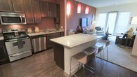 This single-floor space showcases a thoughtfully designed open-concept layout that integrates the kitchen, dining, and living areas into one cohesive environment. The kitchen is a standout feature, equipped with rich dark-stained cabinetry, light granite countertops, and stainless steel appliances including a dishwasher. A large kitchen island with a matching granite surface serves as a functional centerpiece, offering bar-style seating with modern stools — ideal for casual dining or entertaining. Three striking red pendant lights hang above, adding a bold design element and warm ambiance to the space. The flooring throughout features wood-look planks, lending a modern yet warm aesthetic. A deep burgundy/mauve accent wall creates a dramatic visual anchor between the kitchen and the adjacent living area, which is visible in the background and benefits from natural light through what appears to be a large window or sliding door. The dining area sits comfortably between the kitchen and living space, reinforcing the open, connected flow of the layout. Overall, the space balances contemporary design with functional living, creating an inviting and well-appointed interior.