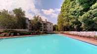 This amenity space offers residents two distinct recreational areas designed for both relaxation and active fitness. The centerpiece is a generously sized outdoor swimming pool with striking turquoise water, set against a backdrop of lush, mature trees that provide natural shade and a serene atmosphere. The pool deck features ample lounge seating arranged along the perimeter, making it an ideal setting for leisure. A white fence encloses the area, adding a classic, clean aesthetic to the outdoor environment.

Adjacent to the pool is a well-appointed fitness center housed in a bright, inviting structure. The gym is divided into multiple zones, including a cardio area lined with large windows that flood the space with natural light and offer views of the surrounding greenery. Equipment includes Cybex cycling bikes, treadmills, and Hoist strength-training machines. A skylight overhead enhances the sense of openness in the strength-training section, where rubber flooring provides a safe and functional workout surface. Additional amenities include stability balls and wall-mounted mirrors that expand the visual space. The neutral gray tones and white trim throughout create a clean, modern aesthetic that complements the well-maintained outdoor surroundings.