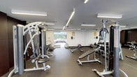 This fitness center offers a thoughtfully designed workout environment that balances functionality with contemporary aesthetics. The space is finished with durable rubber flooring throughout, providing a safe and practical surface suited to a full range of exercise activities.

The strength training area is well-stocked, featuring a multi-station cable machine, a free-weight rack with a graduated dumbbell selection, multiple adjustable weight benches, and additional resistance machines for targeted muscle training. A yoga mat is also available for stretching and floor-based exercises.

The cardio zone is positioned along a floor-to-ceiling glass window wall that frames outdoor views of mature trees and surrounding greenery, creating an energizing atmosphere during workouts. Equipment includes treadmills, elliptical trainers, and stationary bikes.

Linear LED lighting runs across the ceiling, ensuring the entire space is evenly and brightly illuminated. Dark wood-paneled accent walls add a refined, upscale touch that contrasts with the clean white walls. Full-length mirrors are strategically placed to enhance the sense of space and support proper form during exercise. Overall, the fitness center provides a comprehensive and polished environment for a wide variety of training needs.