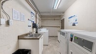 This shared laundry facility is a dedicated, single-level utility space designed to accommodate multiple users efficiently. The room is laid out in a narrow, elongated configuration, with machines positioned along both sides to maximize available floor space.

Along one wall, a row of top-loading washing machines provides ample washing capacity, while the opposite side features a bank of Huebsch commercial-grade dryers equipped with digital displays and card or coin payment systems. Instructional and operational signage is posted prominently throughout the room, offering guidance on machine usage.

A white base cabinet with a stainless steel utility sink is conveniently positioned near the entrance, offering a practical handwashing or pre-treatment station. Additional white cabinetry with countertops provides surface space for sorting or folding laundry.

The flooring consists of neutral-toned tile, and the walls feature a textured finish in a light cream tone. Overhead fluorescent tube lighting ensures the space is well-lit. Exposed plumbing pipes run along the upper walls, reflecting the room's utilitarian character. A single door provides access to and from the facility, contributing to a straightforward, functional layout suited for shared residential use.