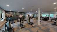 Located at 3000 Shadowood Pkwy SE in Atlanta, GA, this fitness center offers a comprehensive workout facility spread across a single floor. The space is divided into two distinct areas designed to accommodate a variety of exercise needs.

The main workout floor is expansive and features patterned, multi-tone carpet tiles that add visual texture to the space. Structural columns with decorative bases punctuate the floor, and recessed lighting throughout ensures a well-lit environment. Bold teal accent walls contrast with neutral gray surfaces, giving the gym an energetic yet polished aesthetic. This area is outfitted with a full range of cardio equipment including treadmills, ellipticals, and stationary bikes — several featuring integrated display screens — alongside a cable machine, a dumbbell rack, and weight benches. Large windows along one wall invite natural light and provide views of the surrounding greenery.

The secondary room serves as a dedicated studio or group exercise space, finished with warm hardwood-style flooring that sets it apart from the main floor. This room features a wall-mounted flat-screen TV, stability balls, medicine balls, exercise mats, a step platform, and wall-mounted fans. A mirrored door connects it to the main gym, maintaining an open, cohesive feel. Together, the two areas create a versatile fitness facility suited for cardio, strength training, and group or individual exercise sessions.