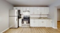 Located at 1150 Vine St in Liverpool, NY, this single-floor unit offers a thoughtfully updated interior with a clean, modern aesthetic throughout. The kitchen is a standout feature, showcasing crisp white shaker cabinetry with brushed nickel hardware, light granite countertops, a subway tile backsplash, and a full suite of stainless steel appliances including a refrigerator, electric range, and over-range ventilation hood. Light wood-tone luxury vinyl flooring runs through the kitchen and living areas, lending warmth and continuity to the open layout. The living area is generously sized, bathed in natural light from a large sliding glass door that opens to a private balcony with outdoor views. A secondary window in the adjacent dining area further enhances the bright, airy feel of the main living space. The bedrooms are finished with plush wall-to-wall carpeting and wide windows that frame wooded outdoor scenery, providing a quiet and comfortable retreat. Baseboard heating is present throughout. The bathroom is a refined space featuring marble-look tile flooring, a tub-shower combination with dramatic dark stone tile surround, and matte black fixtures that add a contemporary contrast. Neutral gray wall tones and clean white trim unify the space with a cohesive, updated look.