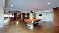 This single-floor recreational space is thoughtfully designed to offer a dynamic and engaging environment for leisure and socializing. At the heart of the room sits a striking billiards table with a rich wood frame, metallic accents, and a bold red felt surface, illuminated by a trio of sleek black pendant lights that draw the eye and define the space. The polished aggregate flooring adds an industrial-chic aesthetic that flows seamlessly throughout the area. A sculptural rope or wire climbing structure anchors one corner of the room, introducing a playful, artistic element that doubles as an interactive feature. Nearby, bar-height seating and accent chairs in vibrant tones create casual gathering spots, while a wall-mounted flat-screen television provides an additional entertainment focal point. Structural columns clad in warm wood tones complement the contemporary design, adding depth and visual interest. Recessed lighting throughout ensures the space is well-lit, while the open layout promotes a sense of spaciousness and easy movement. The overall ambiance is modern and energetic, making this space well-suited for recreation, social events, and community engagement.