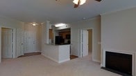 This single-floor residence offers a well-designed open-concept layout that connects the living area and kitchen in a cohesive, functional arrangement. The living room is finished with soft neutral carpeting and features crown molding along the ceiling, a ceiling fan for year-round comfort, and a fireplace that serves as a focal point along one wall. Recessed lighting and flush-mount fixtures provide consistent illumination throughout the space.

The kitchen is partially open to the living area, separated by a raised breakfast bar with a granite countertop — ideal for casual dining or additional workspace. Warm-toned wood cabinetry lines the kitchen walls, complemented by black appliances including a refrigerator and overhead microwave. The kitchen flooring transitions to hard surface material, distinguishing it from the carpeted living space.

Crown molding and clean white baseboards run throughout, lending a polished, traditional character to the interior. Multiple doorways visible from the main living area provide access to additional rooms, including what appears to be a closet with louvered doors and at least one adjacent bedroom or utility space. The neutral color palette of warm beige walls creates a bright and welcoming ambiance throughout this thoughtfully laid-out single-level home.
