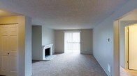 This single-floor space presents a comfortable and functional living area finished in a neutral, understated palette. The main living room is carpeted in a light beige tone that extends throughout, contributing to a warm and cohesive atmosphere. A white brick fireplace anchors one corner of the room, serving as a notable architectural detail that adds character and a traditional focal point to the space.

Natural light enters generously through a large sliding glass door fitted with vertical blinds, which also provides direct access to an outdoor area. This feature brightens the interior and creates a seamless connection between the indoor living space and the exterior. The walls are painted in a soft, neutral gray-white tone that complements the light carpeting and brick fireplace surround.

The ceiling features a textured popcorn finish, consistent with many residential builds of its era. The open layout of the main living area allows for flexible furniture arrangement. Overall, the space presents a clean, move-in ready environment with classic residential details and practical design elements throughout.