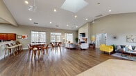 Located at 101 Carden Dr in Mebane, NC, this single-story community clubhouse offers a well-appointed gathering space designed for both relaxation and activity. The entrance lobby makes a strong first impression with bold deep-green accent walls, large glass double doors, tile flooring, and ample natural light from surrounding windows. The expansive main lounge features soaring vaulted ceilings, rich hardwood-style flooring, and a striking fireplace with a wood-trimmed mantle topped by a wall-mounted television. Comfortable seating arrangements, vibrant accent chairs, and curated artwork create an inviting, modern atmosphere throughout. A skylight adds to the bright, airy feel of the space. The dining and social area includes multiple high-top tables and bar-style seating set beneath vaulted ceilings, adjacent to a full kitchen equipped with wood cabinetry, black appliances, tile flooring, and a pass-through counter. A dedicated package locker room provides a secure, convenient solution for parcel deliveries. The fitness center is generously equipped with treadmills, ellipticals, strength-training machines, and free weights, complemented by teal accent walls, ceiling fans, skylights, floor-to-ceiling mirrors, and carpeted flooring. Together, these amenities create a functional and welcoming community hub.