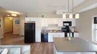 This single-floor residence offers a thoughtfully designed layout that balances functionality and comfort throughout its key living spaces. The open-concept kitchen is a standout feature, equipped with white shaker cabinetry, grey countertops, a double-basin sink, and a full suite of black appliances including a refrigerator, electric range, built-in microwave, and dishwasher. Track lighting and a peninsula with pendant lights add both practicality and visual appeal. Adjacent to the kitchen, the dining area accommodates a round table and benefits from a large window that invites natural light and outdoor views. The living room is generously sized with warm neutral tones, carpet flooring, and sliding glass doors that open directly onto a private balcony, extending the living space outdoors. Two well-proportioned bedrooms each feature plush carpet, wood-panel headboards, and walk-in or reach-in closet access. The two bathrooms are finished with white vanities, large mirrors, multi-bulb lighting fixtures, and tub-shower combinations. Throughout the residence, wood-look flooring in the main living areas and warm grey wall tones create a cohesive, modern aesthetic.