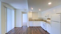 This single-floor space showcases a well-appointed kitchen that serves as a central feature of the layout. The kitchen is outfitted with crisp white shaker-style cabinetry that extends to the upper walls, providing generous storage. Granite countertops in warm, neutral tones add a touch of elegance and durability to the workspace. White appliances, including a gas range, built-in microwave, dishwasher, and refrigerator, are seamlessly integrated into the cabinetry for a cohesive look. A single-basin sink is positioned beneath upper cabinets, offering a functional and tidy layout. Rich, dark hardwood-style flooring runs throughout the space, creating a striking contrast against the white cabinetry and light-colored walls. Recessed lighting in the ceiling provides bright, even illumination across the kitchen and adjacent areas. The walls are finished in a clean, neutral white, enhancing the sense of openness and light. A doorway visible from the kitchen leads to an additional area of the unit, suggesting a practical and connected floor plan. Natural light filters in, further contributing to the airy, welcoming ambiance of the space.