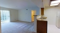This single-floor residential space offers a functional and open layout that maximizes the sense of space and comfort. The living area is generously sized, featuring wall-to-wall carpeting that adds warmth underfoot and a vaulted ceiling that creates an airy, expansive feel overhead. Neutral wall tones throughout provide a clean, versatile backdrop.

The kitchen is positioned adjacent to the living area and is defined by a half-wall breakfast bar that serves as a natural dividing element while maintaining the open flow between spaces. The kitchen is equipped with warm wood-toned cabinetry, a double sink set into a light-colored countertop, and ample cabinet storage. A doorway visible from the main living area leads to additional rooms within the unit, suggesting a well-connected and practical floor plan.

Natural light filters into the space, complementing the light color palette of the walls and ceiling. Ceiling vents indicate a central HVAC system for year-round climate control. Overall, the space presents a straightforward and livable residential environment, well-suited for everyday living with its cohesive design and efficient use of the single-floor layout.