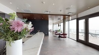 This elegantly designed single-floor amenity space offers a seamless blend of modern architecture and refined interiors. The open-plan layout connects a sleek, fully equipped kitchen to a spacious lounge area, creating a cohesive and functional environment. The kitchen features a striking marble backsplash, rich dark wood cabinetry, built-in stainless steel appliances including a wall oven and beverage cooler, and a generously sized island with a clean white countertop — ideal for both casual gatherings and catered events. Adjacent to the kitchen, a large reception-style bar counter with a marble surface and warm wood-slat ceiling detail adds an upscale hospitality feel. The lounge area is anchored by a sculptural pendant light fixture suspended above a central table, surrounded by deep burgundy leather chairs and contemporary seating arrangements. Expansive floor-to-ceiling glazing floods the interior with abundant natural light while framing dramatic, panoramic city skyline views. A set of French doors opens directly onto a spacious outdoor terrace, extending the living area and offering an elevated vantage point over the surrounding urban landscape. Exposed concrete columns and accent walls provide an industrial contrast to the warm wood ceiling panels and light wood-tone flooring throughout, resulting in a space that is both visually dynamic and inviting.