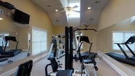 This single-floor home fitness space is thoughtfully designed to support a full range of exercise routines. The room is anchored by a multi-station cable and weight machine, offering a variety of strength training options, complemented by additional cardio and resistance equipment. The space is finished with neutral-toned walls and light carpeting, creating a clean and motivating workout environment. Vaulted ceilings rise dramatically overhead, enhancing the sense of openness and allowing for comfortable use of tall equipment. A clerestory window positioned near the ceiling peak floods the room with natural light, while a ceiling fan ensures proper air circulation during intense sessions. Recessed lighting throughout provides consistent, even illumination. Multiple windows fitted with blinds line the walls, offering additional natural light and the option for privacy. A large wall mirror reflects the space, making the room feel more expansive while also serving as a practical tool for monitoring form during exercise. The overall ambiance is functional yet well-appointed, delivering a private gym experience within a comfortable residential setting.