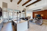 Located at 9125 Highway 6 N in Houston, TX, this thoughtfully designed single-floor community space offers a generous, open-concept layout that seamlessly blends comfort, functionality, and modern style. Soaring ceilings with exposed dark wood beams create a dramatic architectural statement throughout, complemented by light wood-look flooring that ties the space together cohesively.

The main living area features a plush seating arrangement centered on a bold abstract area rug, offering a welcoming lounge setting. A striking textured cork accent wall serves as a focal backdrop for a large flat-screen TV and a sleek media console, adding warmth and visual depth. A designer pendant chandelier in brass anchors the dining zone, where a substantial dark wood table and mixed seating provide an elegant gathering spot beneath a tray ceiling finished in deep navy blue.

A vertical wood slat room divider artfully defines zones without closing off the open floor plan. The kitchen-style refreshment bar, outfitted with counter seating, pendant lighting, and large French doors that flood the area with natural light, adds a social hub to the space. A dedicated co-working nook features a built-in desk with computer stations set against a decorative patterned wallcovering. The recreation area includes a foosball table, rounding out the space's versatile, amenity-rich character.