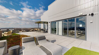 This expansive rooftop terrace is a thoughtfully designed outdoor amenity space that combines contemporary architecture with functional, inviting outdoor living areas. The building's exterior is clad in sleek vertical metal paneling, complemented by floor-to-ceiling glass walls that blur the boundary between interior and exterior spaces.

The terrace is divided into several distinct zones. A sun lounge area features wicker chaise lounges positioned on large-format porcelain pavers, ideal for relaxing under an open sky. Adjacent to this is a casual gathering zone with sculptural lounge chairs and a round table set atop a lush artificial turf lawn, creating a soft, garden-like atmosphere. A separate seating area offers a modular outdoor sofa alongside dedicated game space, adding a recreational element to the rooftop experience.

An outdoor kitchen and dining area, sheltered by a pergola, provides a dedicated space for al fresco entertaining. Throughout the terrace, brick and masonry raised planters incorporate trees and plantings, adding greenery and seasonal interest to the elevated setting.

One of the terrace's most compelling features is its living green roof border, a vibrant band of low-growing ground cover that lines the perimeter walkway and transitions to panoramic views of the surrounding city skyline and open landscape. Glass and metal railings preserve these sightlines while maintaining an open, airy feel across the entire rooftop.