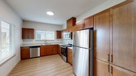 This single-floor unit offers a thoughtfully designed layout with a clean, modern aesthetic throughout. The kitchen serves as a functional centerpiece, featuring warm medium-toned wood cabinetry extending to the upper walls for generous storage capacity. A full suite of stainless steel appliances includes a refrigerator, electric range, built-in microwave, and dishwasher. Light-toned countertops complement the cabinetry, while a double-basin sink is positioned beneath a window fitted with horizontal blinds, allowing natural light into the space. Light wood-style vinyl plank flooring runs throughout the kitchen and living areas, contributing to a bright, cohesive feel.

The living area is open and airy, with neutral white walls and a window with blinds that brings in additional natural light. A wall-mounted HVAC unit provides climate control. The bedrooms feature neutral carpeting and generous wall space, with windows offering views of the surrounding neighborhood. The bathroom is well-appointed with a single-basin vanity, wood cabinetry with brushed nickel hardware, light quartz-style countertops, and a separate shower area. In-unit laundry connections are also present within the bathroom space.