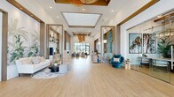 Located in Clermont, Florida, this single-floor clubhouse spans a generously sized footprint and showcases a refined, coastal-modern design throughout. The grand entry lobby immediately impresses with soaring ceilings, warm woven pendant light fixtures, and large-format light wood-toned tile flooring that runs seamlessly across the space. Tall floor-to-ceiling windows and glass entry doors flood the interior with abundant natural light while offering views of the surrounding Florida landscape.

The main lounge area features elegant gold-framed architectural partitions, a built-in fireplace with a sleek stone surround, and curated seating arrangements accented by teal, blue, and neutral tones. A dedicated beverage and refreshment station is tucked into an alcove alongside open shelving and a wine refrigerator. A separate lounge nook showcases a dramatic teal upholstered accent wall with tropical botanical mural wallpaper and plush white sofas, creating a relaxed yet stylish retreat.

Private office or meeting rooms are enclosed by full-height sliding glass doors with modern metal hardware, offering both transparency and flexibility. The hallway leads to additional functional areas, all maintaining the same cohesive palette of warm woods, white walls, and gold accents. The overall ambiance is polished, welcoming, and distinctly contemporary with a Florida-inspired flair.