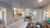 This apartment showcases an open floor plan that seamlessly integrates the kitchen, dining, and living areas. The kitchen boasts white cabinetry and stainless steel appliances, complemented by a breakfast bar. Adjacent is a chic dining area with a round table and modern chairs. The living room features a plush tufted sofa in light blue, centered on a zebra-print rug. Hardwood floors run throughout, while recessed lighting and pendant fixtures provide ample illumination. A geometric wall art piece adds a pop of color, enhancing the space's contemporary ambiance.